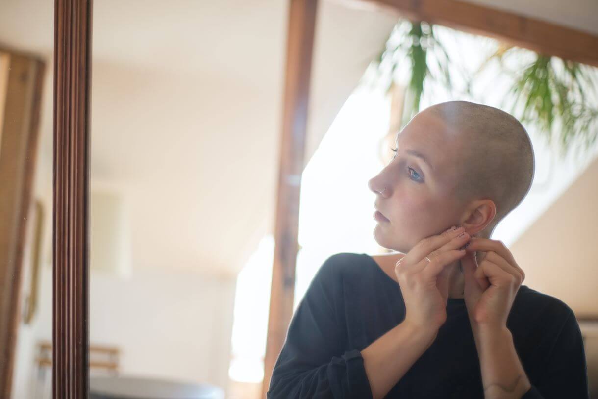 mulher careca colocando brincos em frente ao espelho, representando o autocuidado e autoestima.